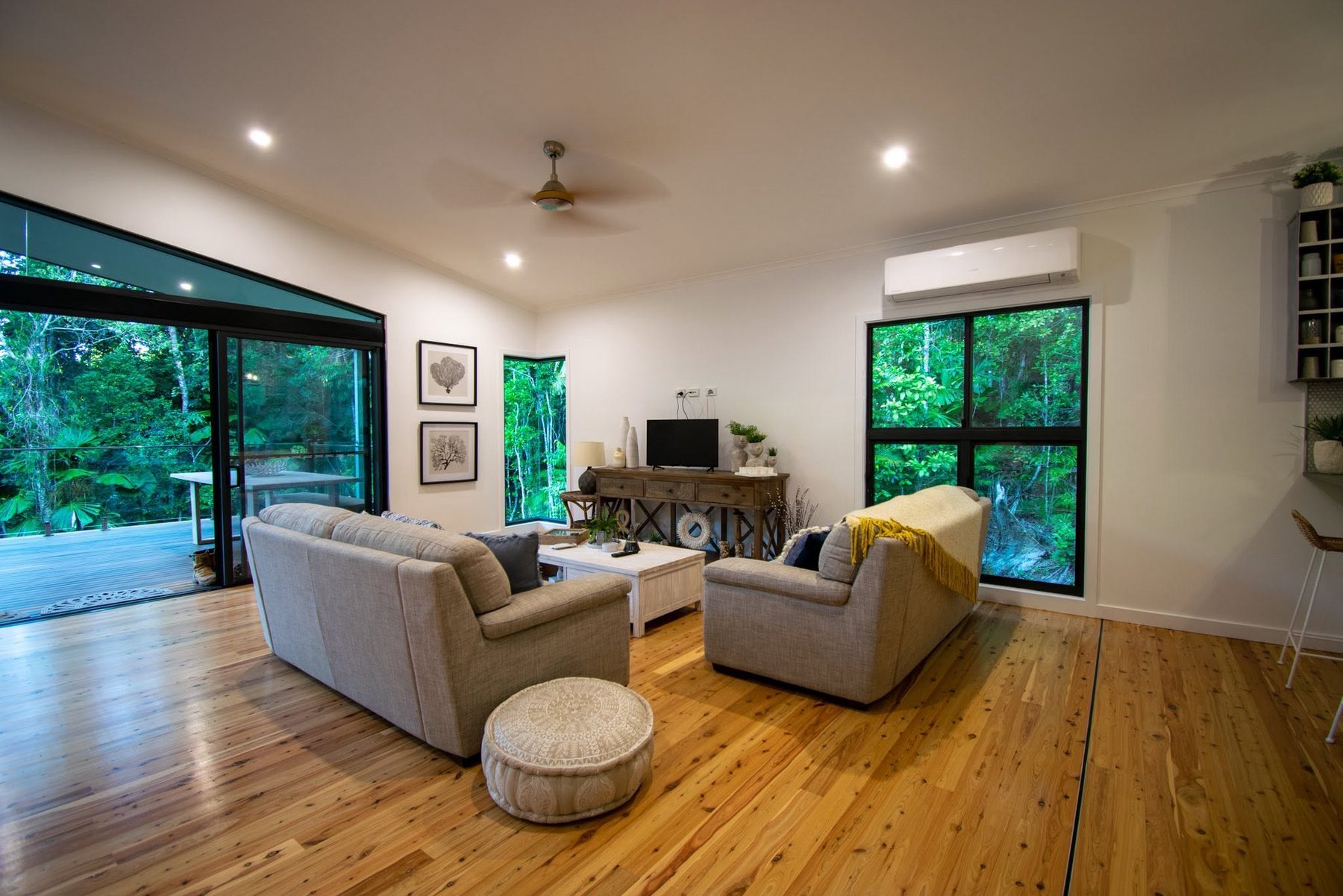 A living room with a ceiling fan, a split system and downlights, rainforest through the glass