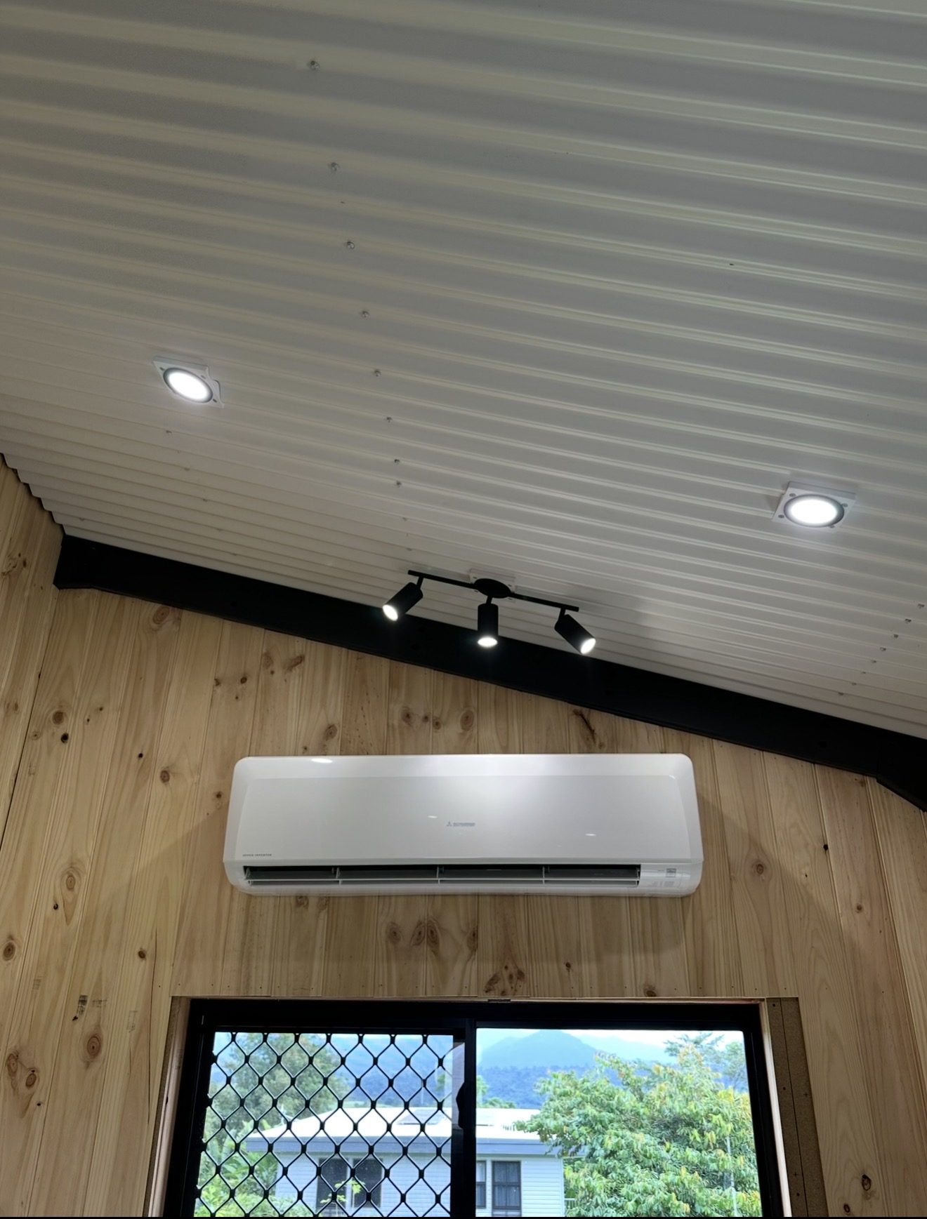 A split system head, track lighting and downlights in a timber lined room looking out at the ranges